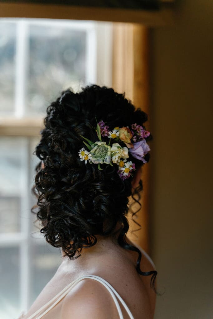 Brunette woman with curly updo adorned with fresh wildflowers, styled for a photoshoot in natural light near a window.