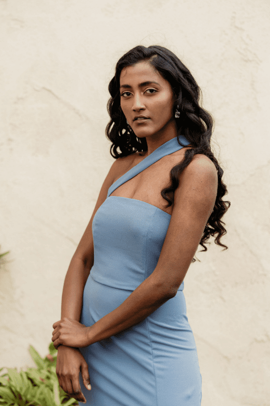 Woman with dark wavy hair and natural glam makeup poses confidently in a light blue one-shoulder dress against a neutral stucco wall