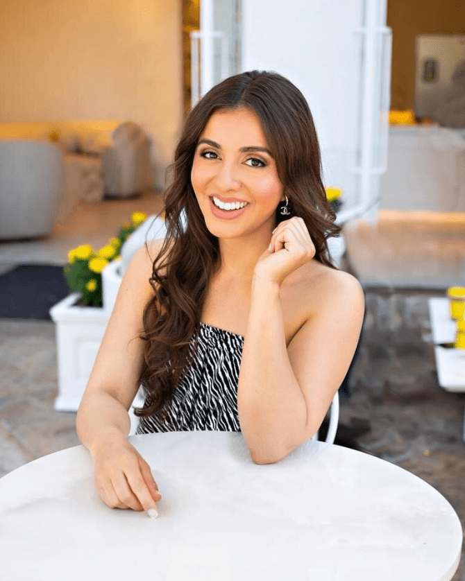 Woman with long brunette hair and soft makeup smiling at the camera while seated at a white table, with yellow flowers in the background.