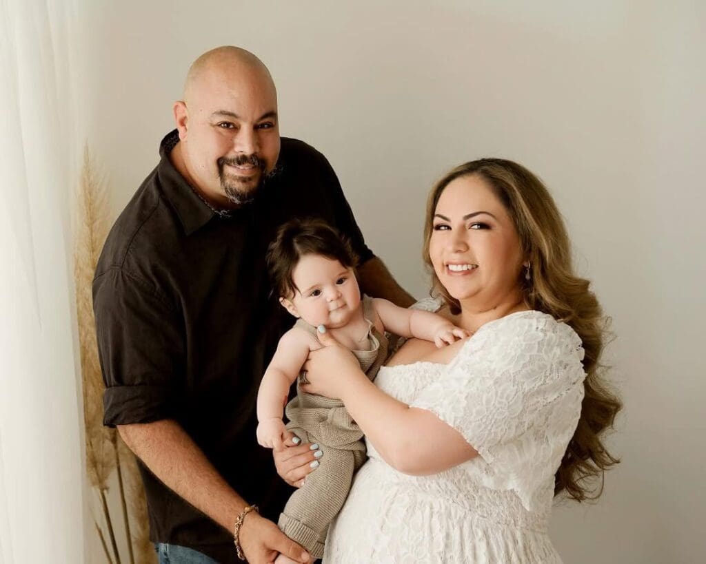 A new family of three smiling in natural light, with mom in a lace dress holding their baby and dad by her side.