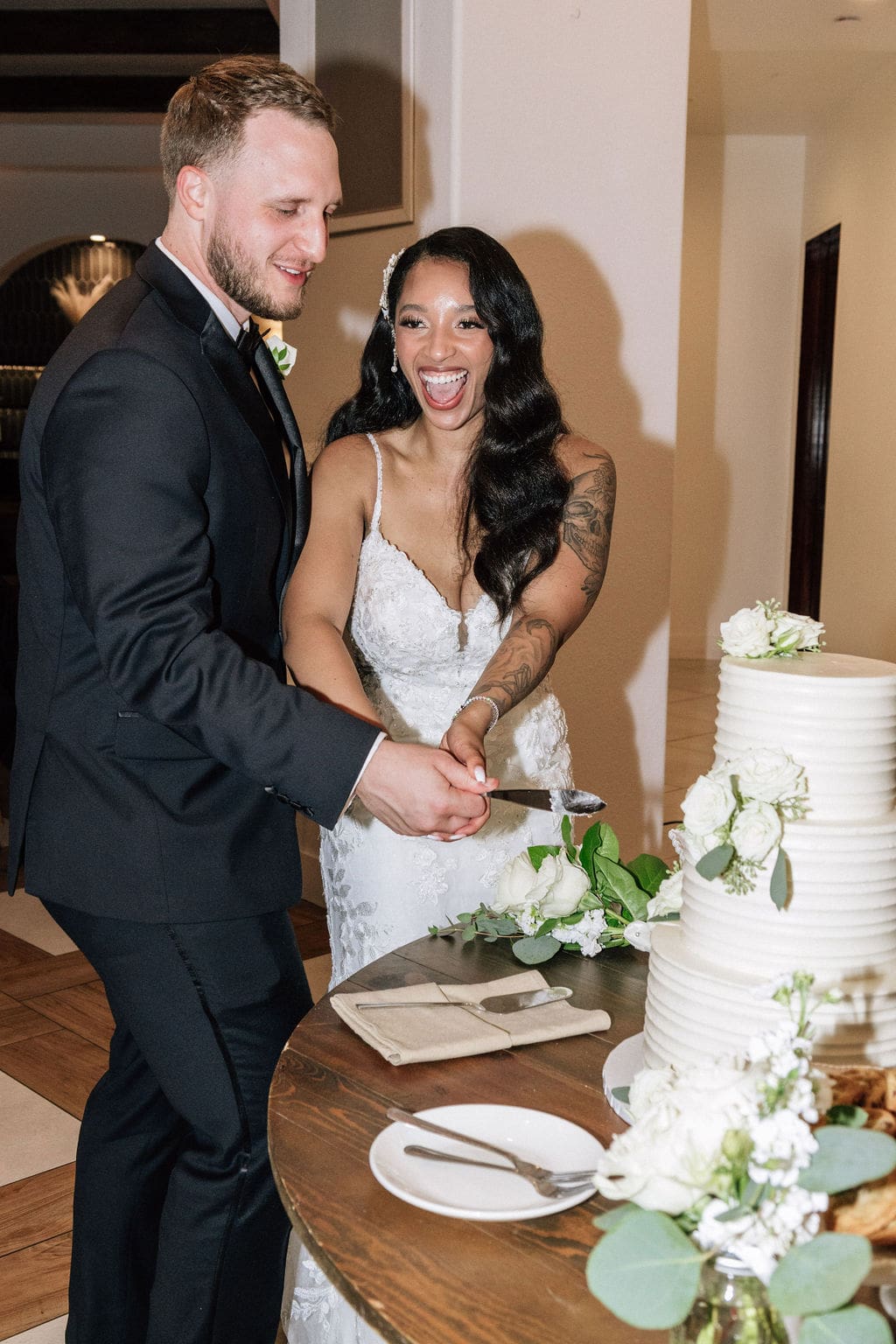 Bride with light brown skin tone with long glamorous waves in her hair, laughing with groom while cutting three-tiered white wedding cake.