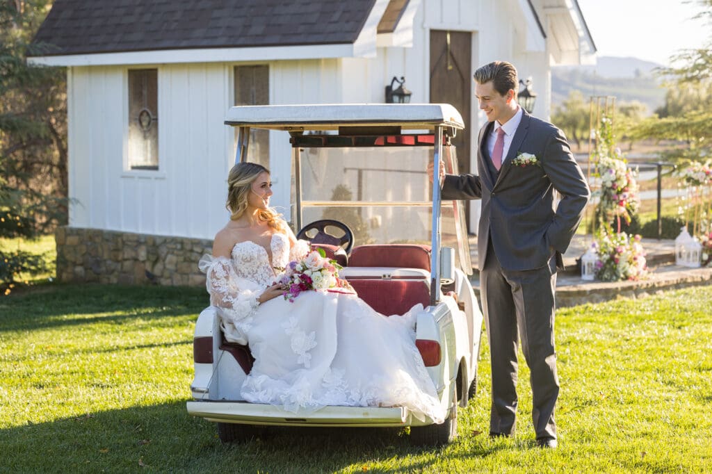 Bride sitting in a golf cart with her bouquet, groom standing beside, both smiling in front of chapel.
