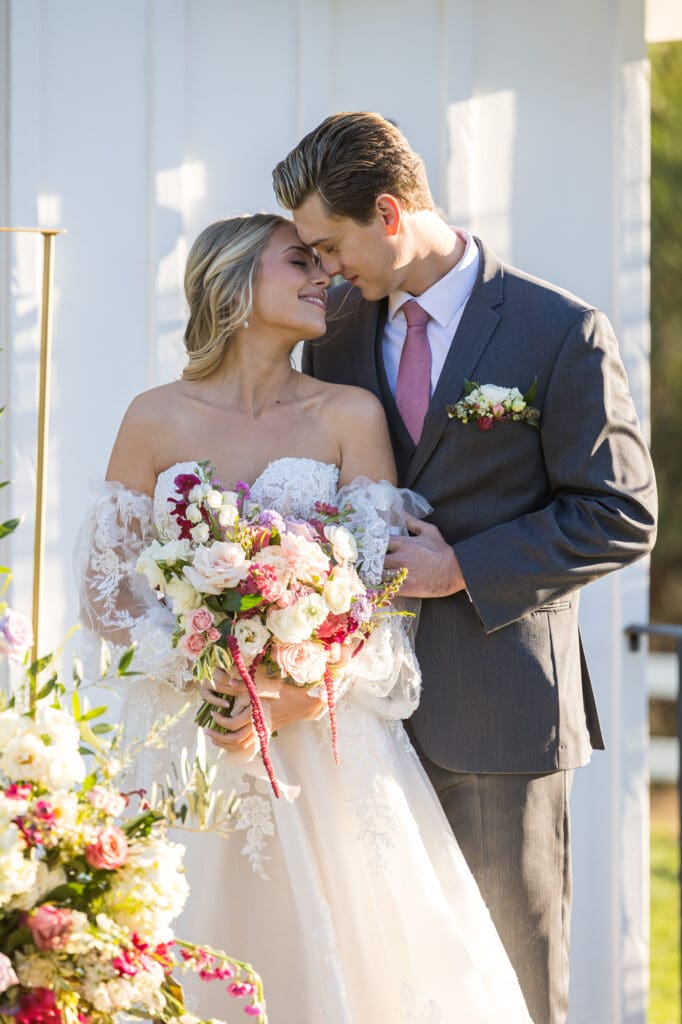 Bride and groom share a tender forehead touch outside the chapel, bride holding bouquet of pink and white blooms.