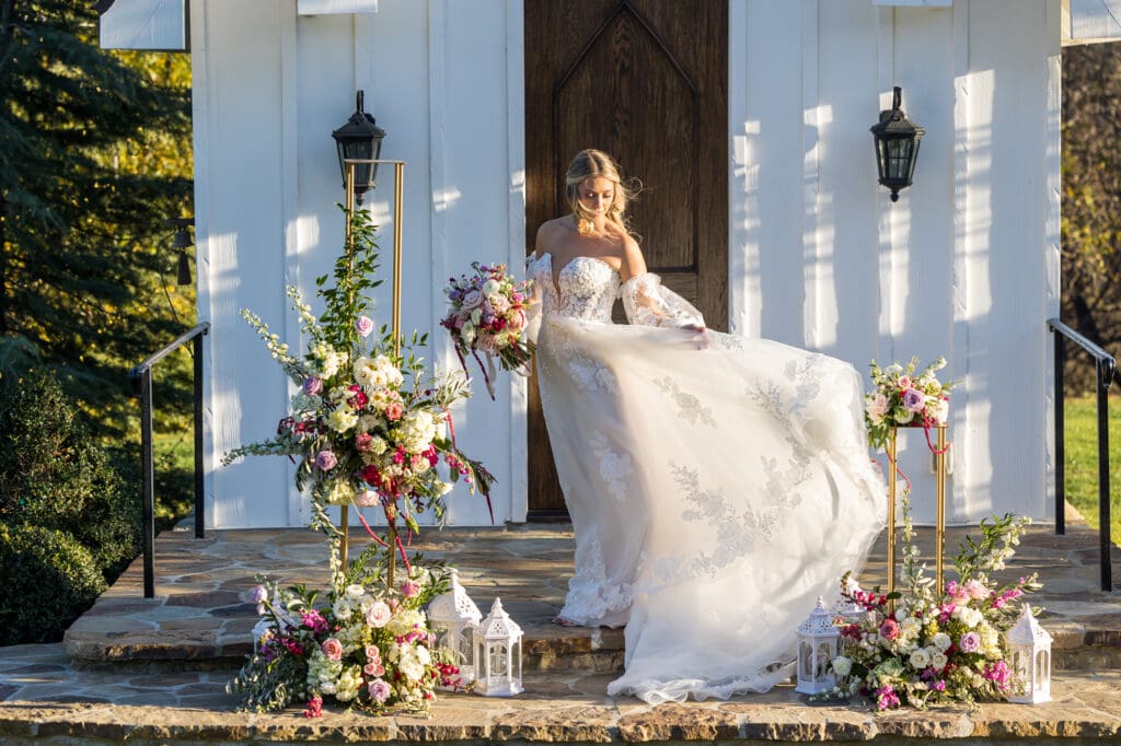 Bride on chapel steps lifting her lace gown, surrounded by floral arrangements and white lantern accents.