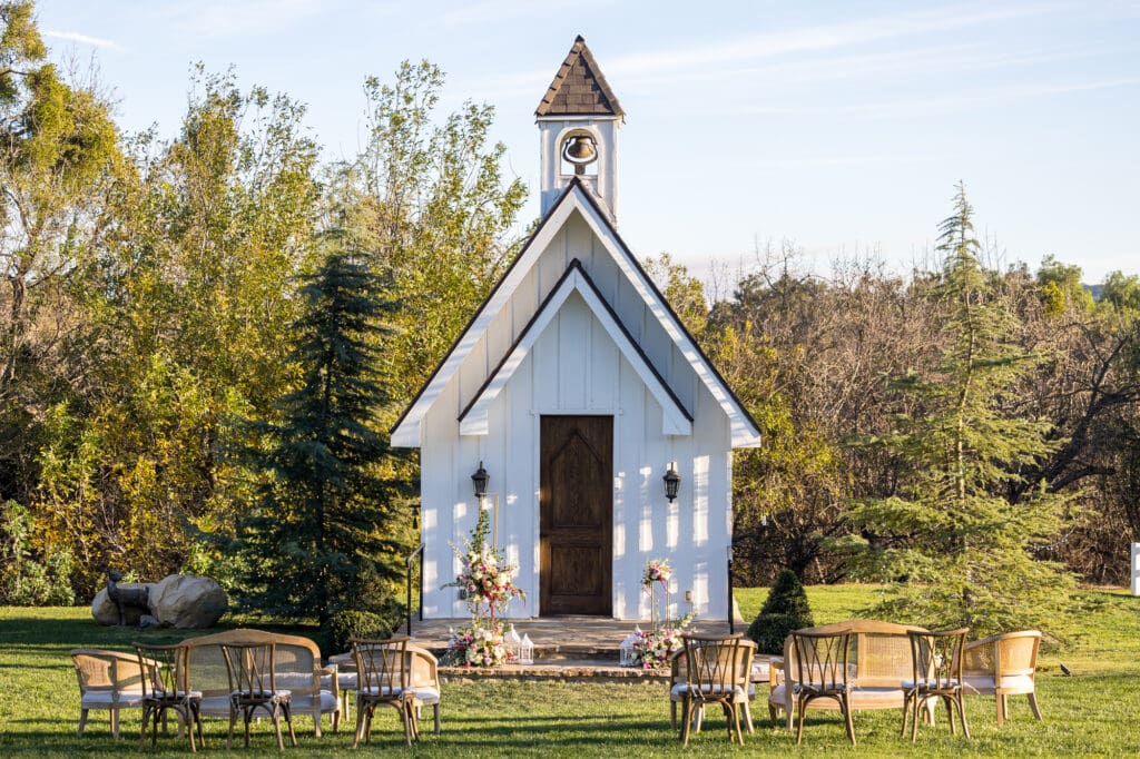 Ceremony setup with white chapel framed by trees, floral displays, and elegant wooden chairs arranged for guests.