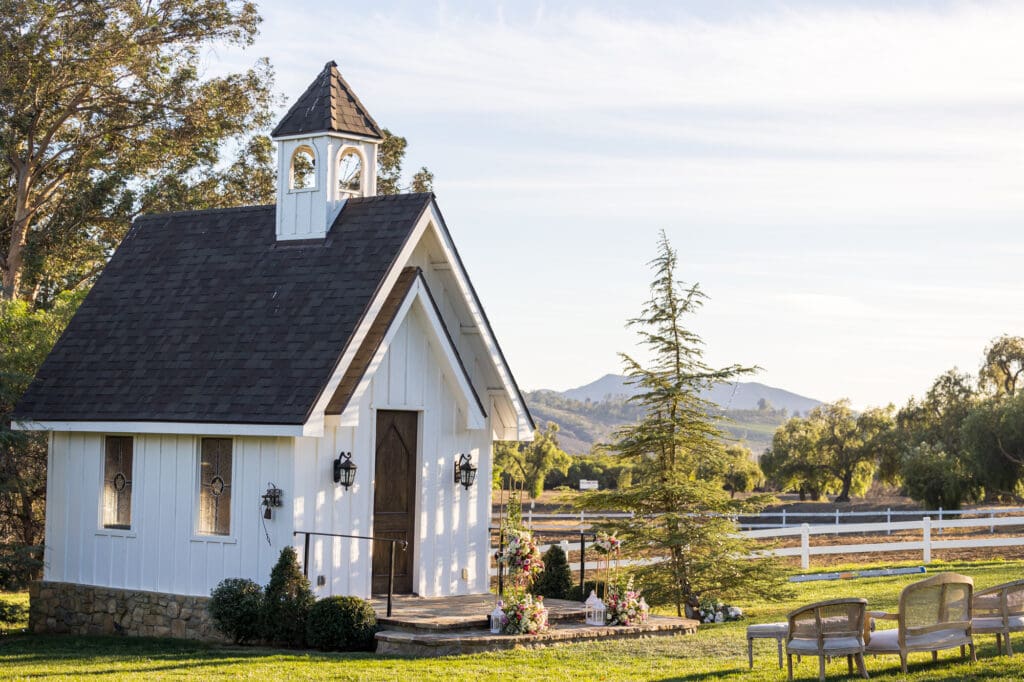 Quaint white chapel with stone steps, rustic lanterns, and floral arrangements, framed by trees and mountain views in the distance.