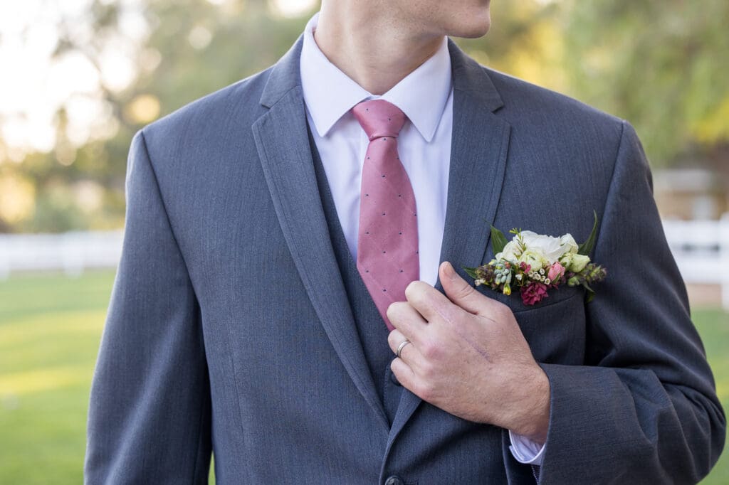 Groom in a gray suit and pink tie, adjusting his lapel with a boutonniere of white and pink flowers.