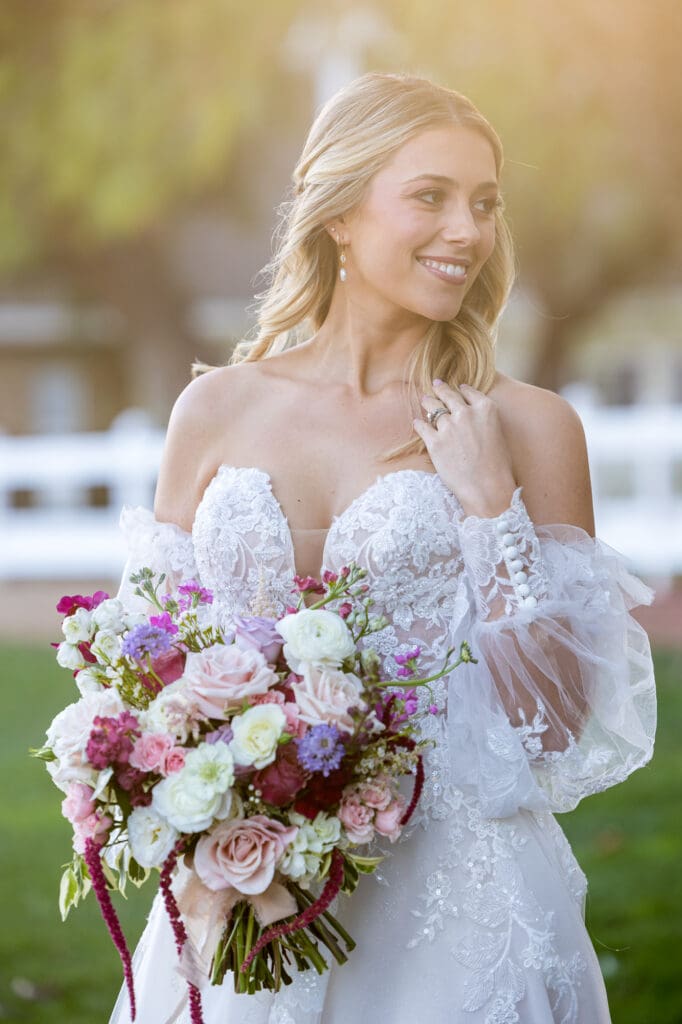 Bride in lace off-the-shoulder gown smiling softly while holding a colorful bouquet of roses and ranunculus.