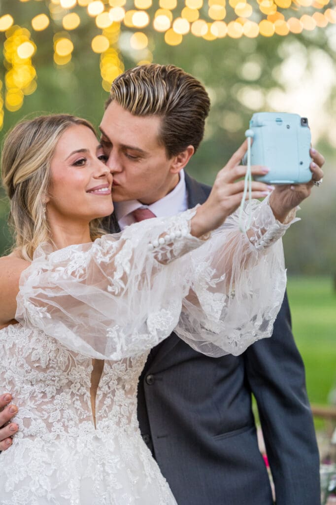 Bride and groom take a Polaroid selfie, groom kissing bride's cheek under twinkling lights.