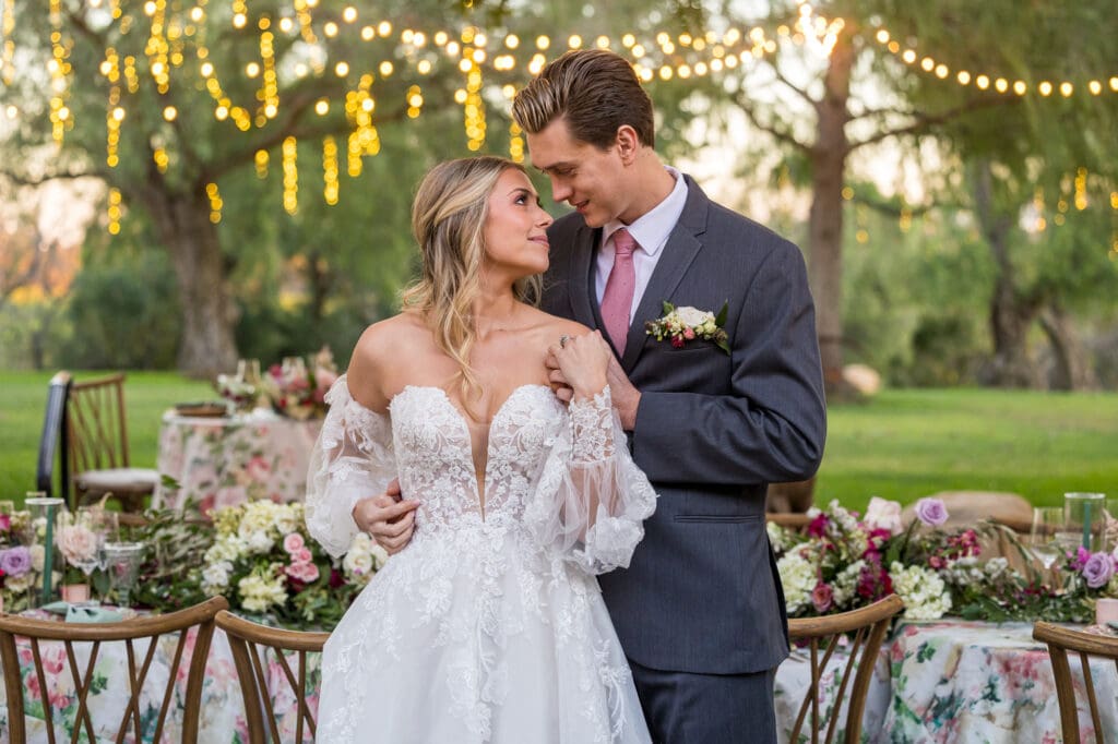 Bride and groom stand close together under string lights, gazing into each other's eyes with floral reception tables behind.