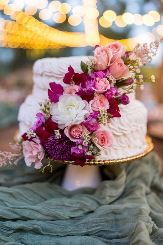 White wedding cake decorated with cascading pink and magenta roses, with glowing string lights win the background.