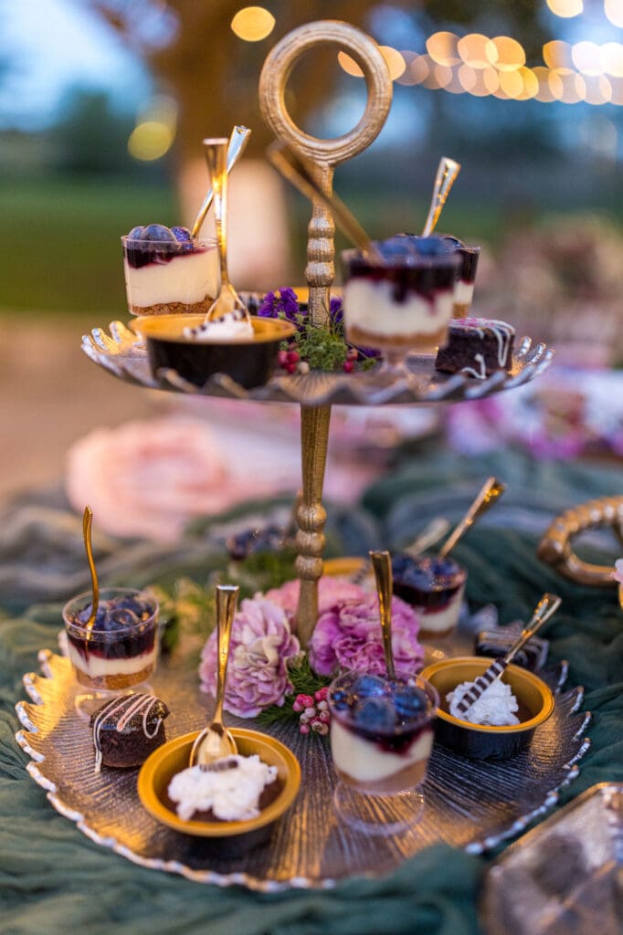 Tiered dessert stand with mini cheesecakes, chocolate mousse, and floral accents, styled with golden utensils.