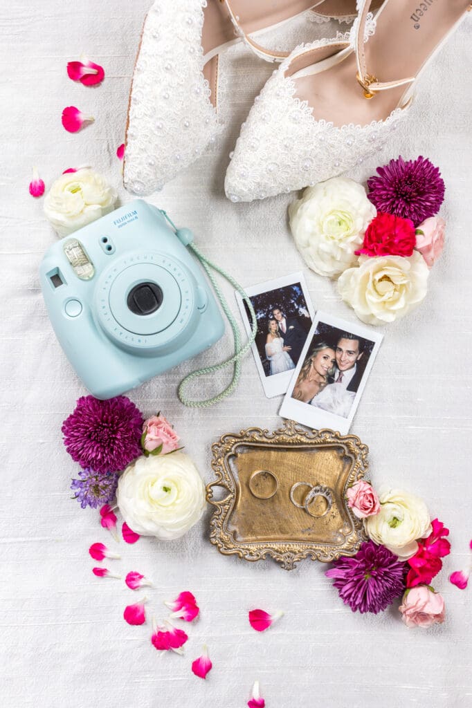 Flatlay of wedding details: lace heels, Instax camera, Polaroid snapshots, wedding rings on vintage tray, and scattered petals.