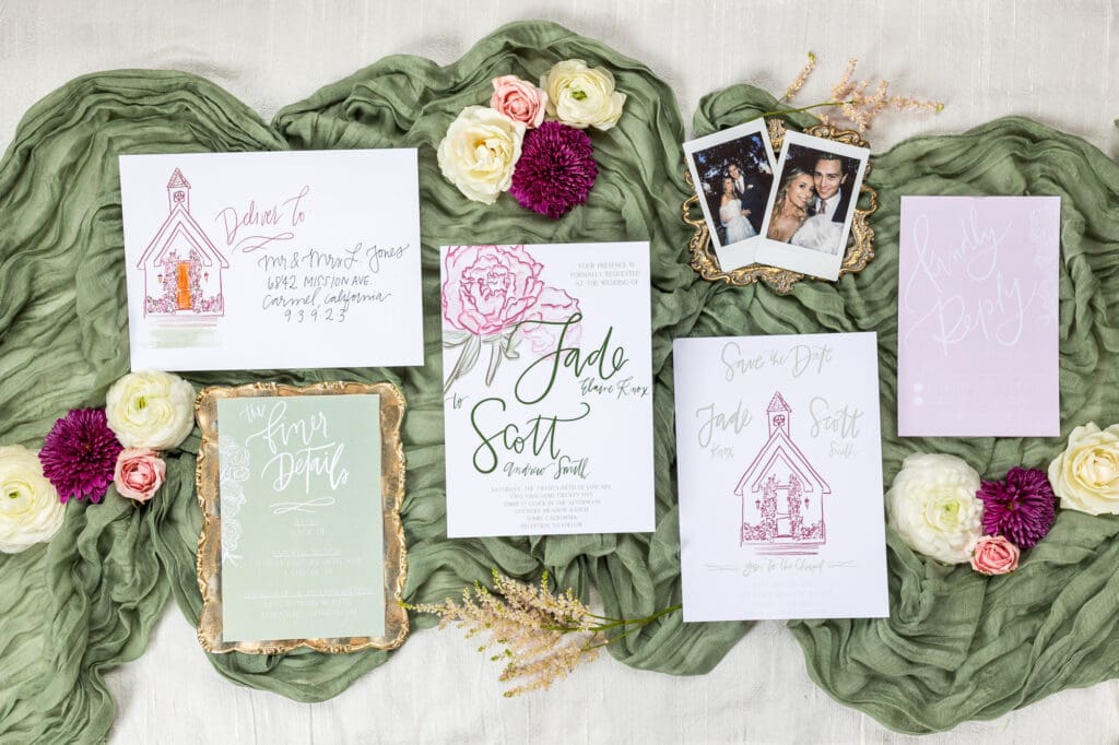 Styled wedding stationary suite with floral illustrations, Polaroids, and calligraphy, laid on green chiffon with blooms.