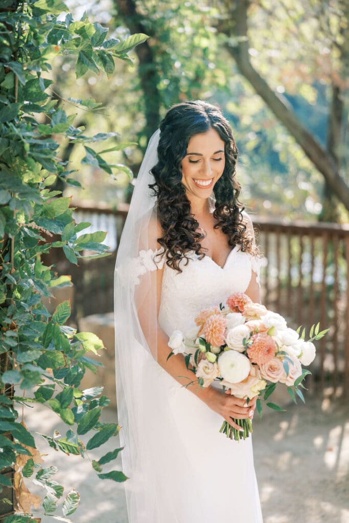 Bride with long, defined curls wearing an off-shoulder gown and veil, styled by Pacheco Beauty for a romantic outdoor wedding